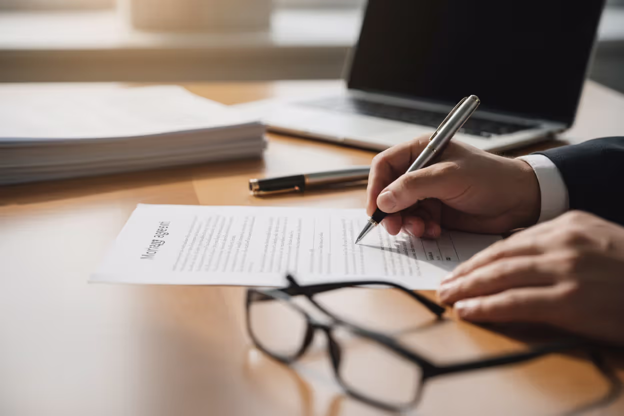 Close-up of a person signing mortgage documents at a desk with glasses and a pen nearby in soft natural lighting
